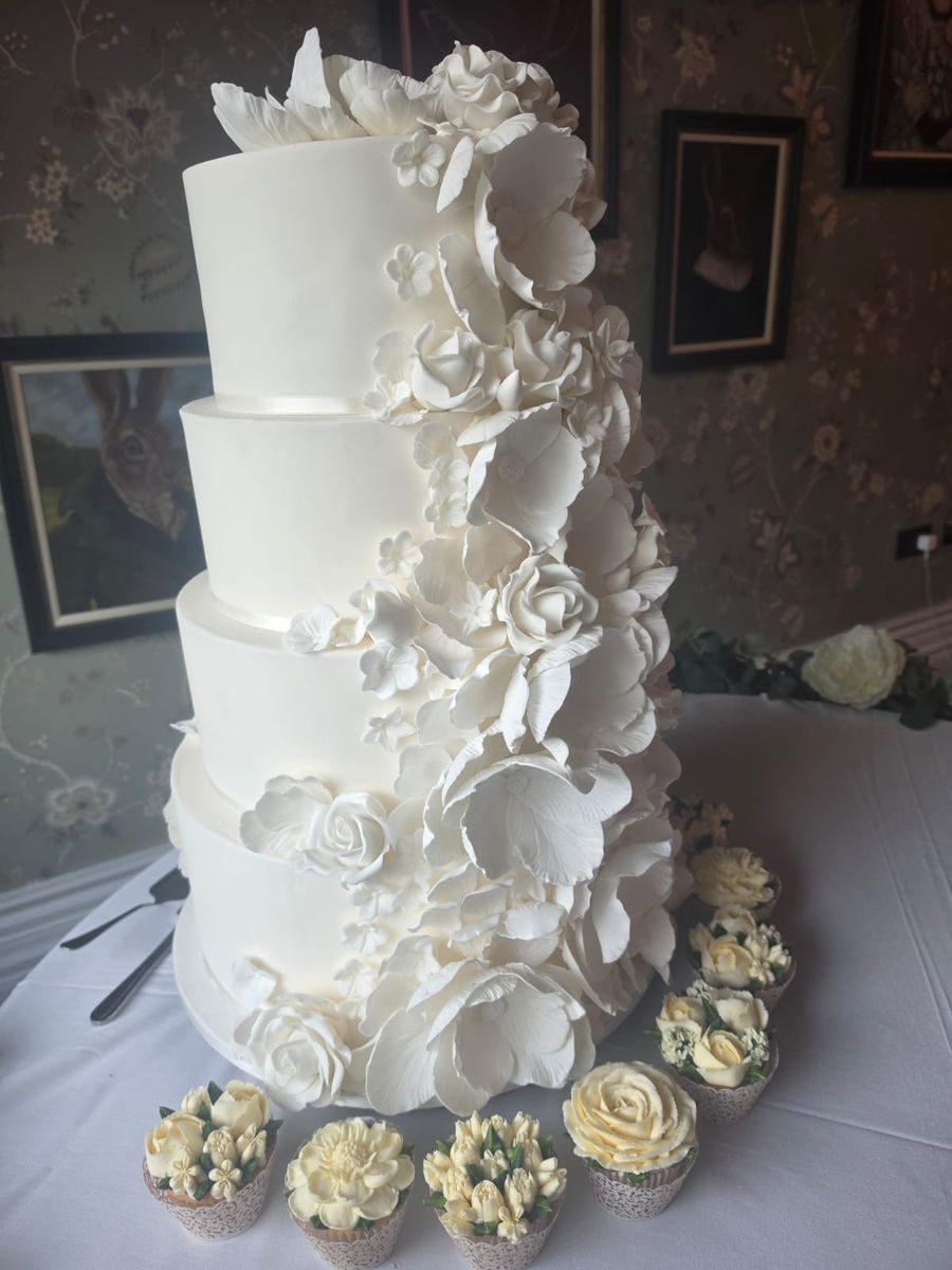 Four tier white cake with a cascade of sugar flowers and cupcakes