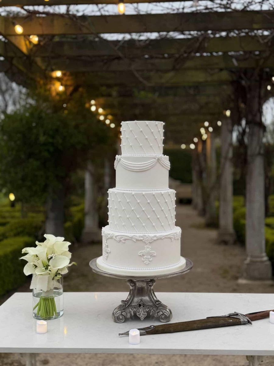 A four tier white Busy Whisk wedding cake with quilted pearl detail under a lit garden pergola